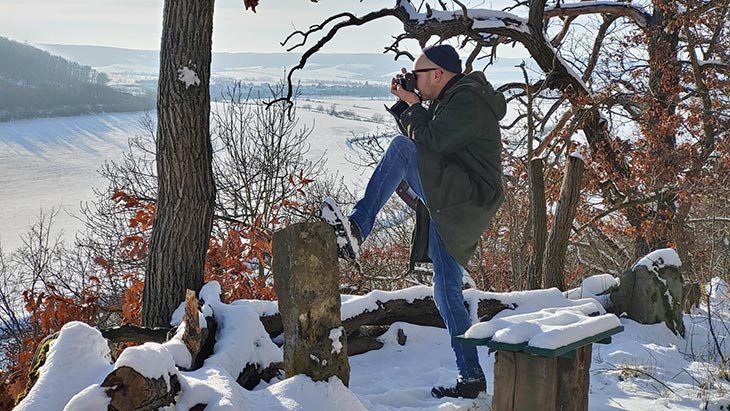 Mann steht mit Kamera in einer Winterlandschaft und stützt seinen Fuß auf einen Stein.