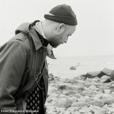 Thomas Raatz steht an einer Küste, Steine und das Meer im Hintergrund.